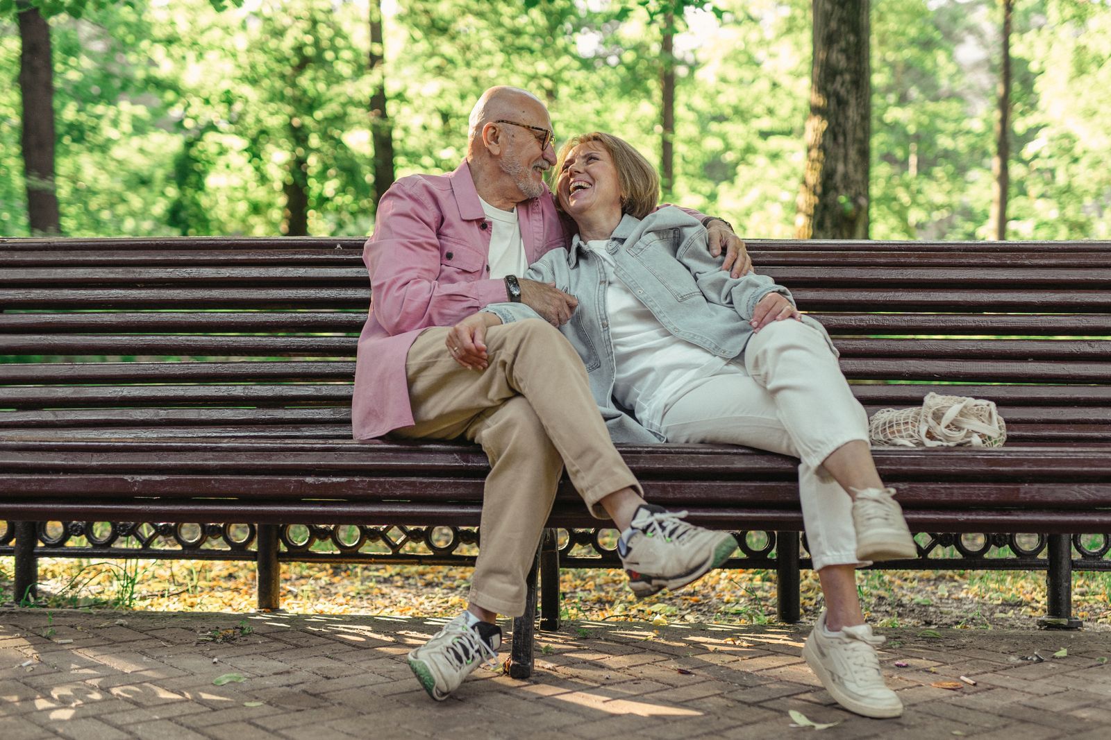 An older couple sitting close together on a wooden bench in a green park, laughing together in the sunshine