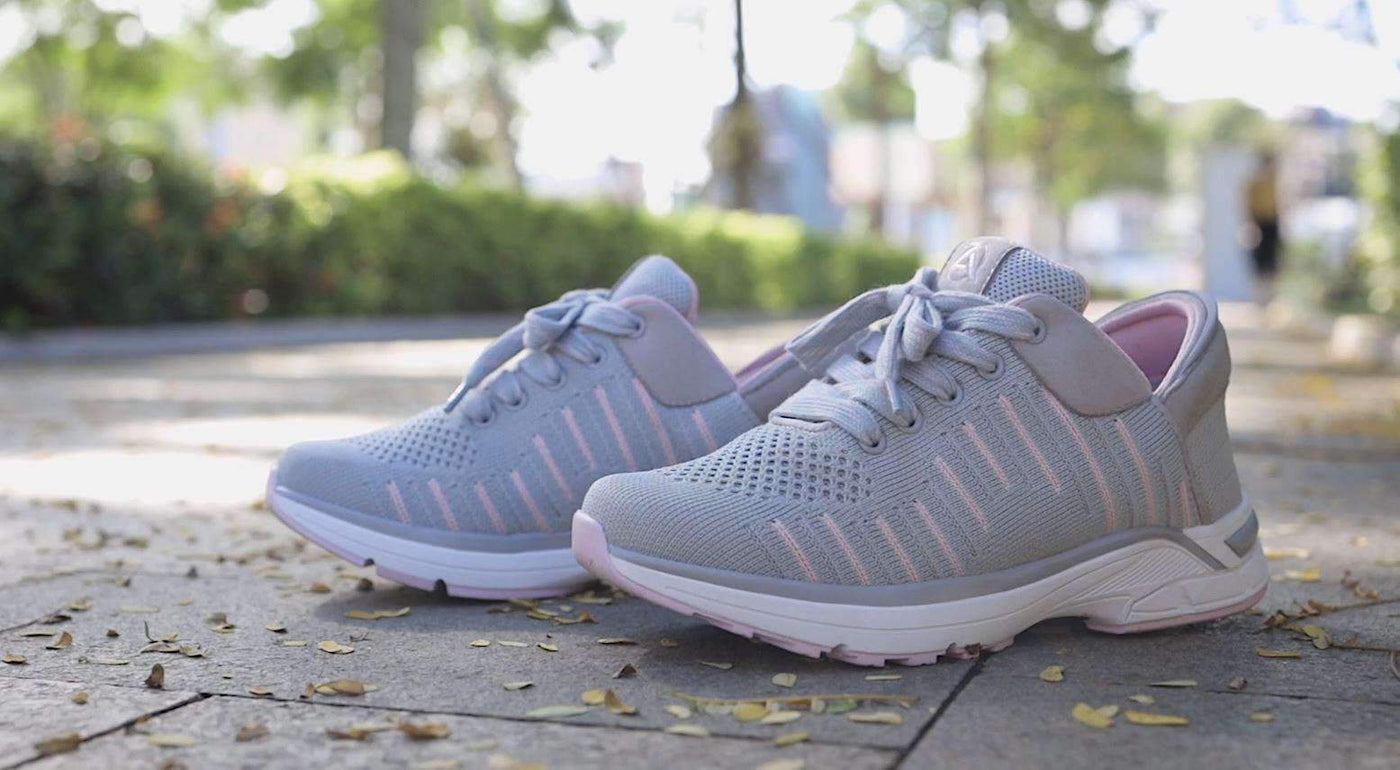 A pair of grey and pink knit Zeba sneakers standing on a paved path, with blurred greenery and fallen leaves behind them
